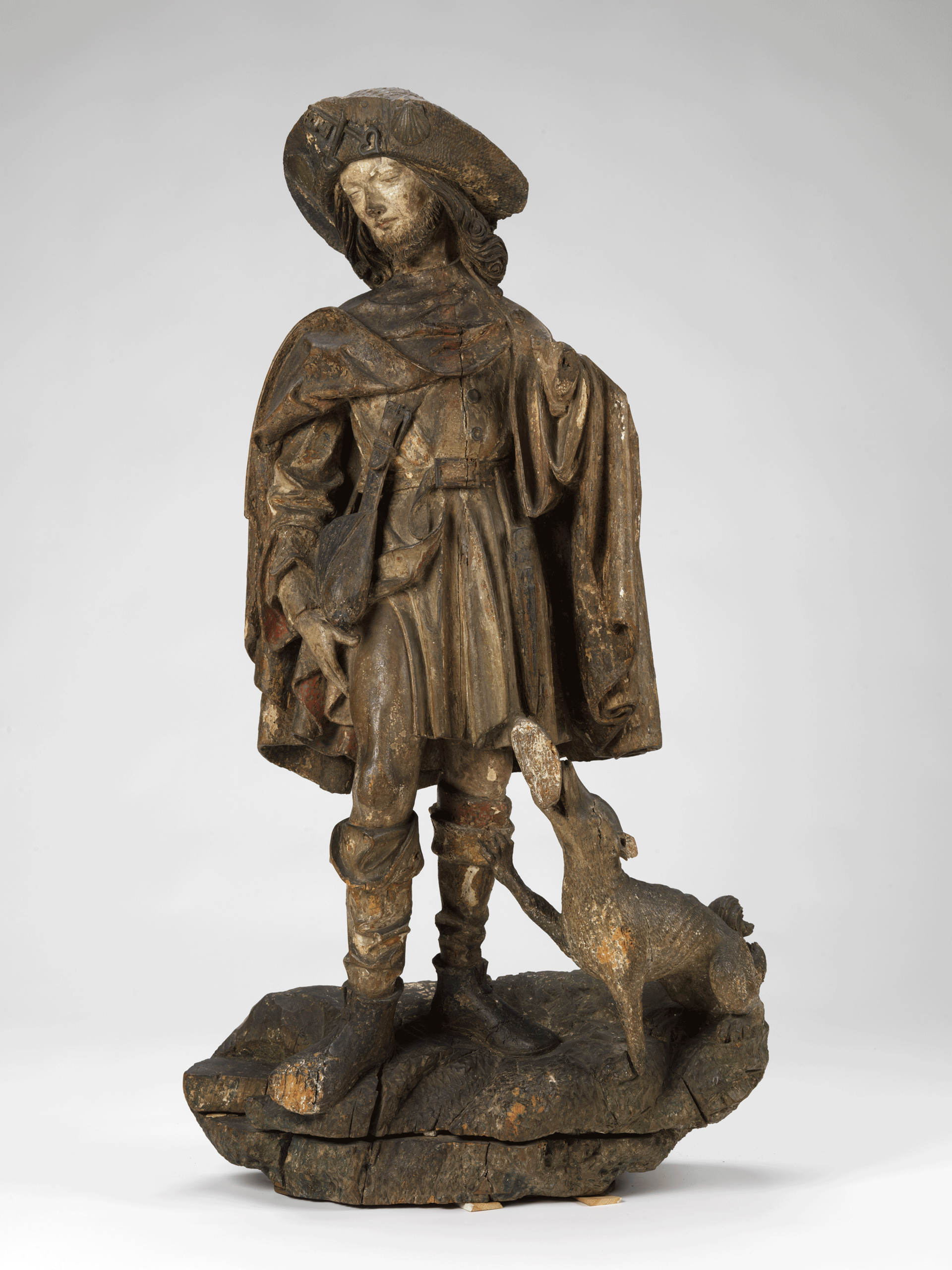 Dark-brown sculpture of a standing man wearing a cape, hat, and boots. He looks to the side, hand on his thigh. A dog at his feet looks up at him.