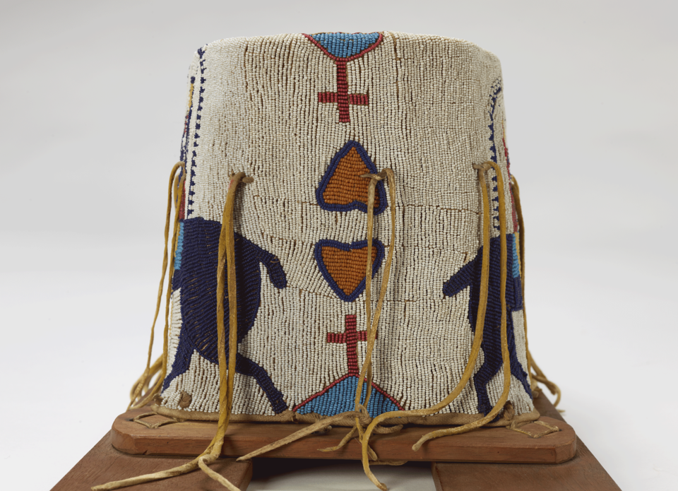 Detail of beaded object with leather ties hanging down and wood supports at bottom. White background beaded with pink and blue hearts and red and blue crosses.  