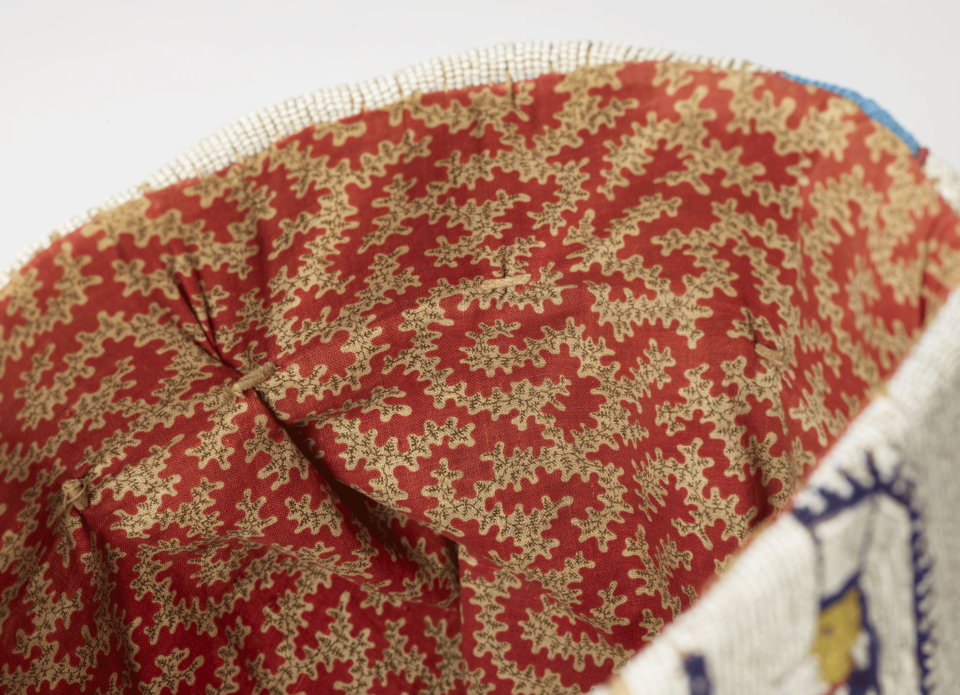 Interior detail of an object, covered in a dense print of cream flowers on a red background. The outer edge of the object is beaded in blue, white, and yellow.